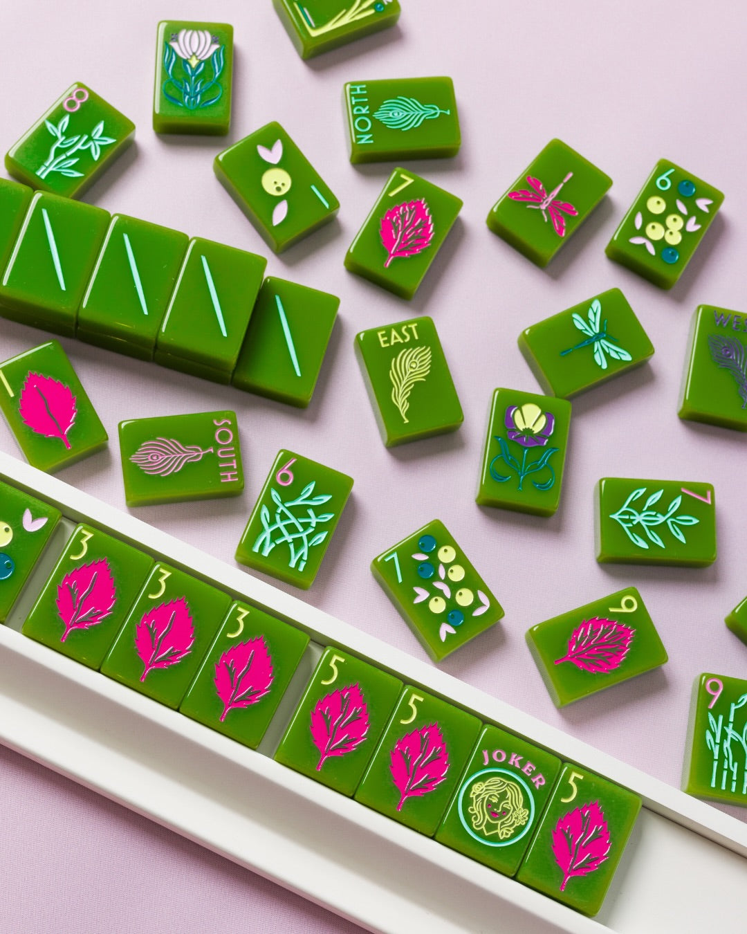 Green Mahjong tiles with colorful Chinese character and floral designs, arranged on a light pink background with a white tray holding several tiles at The Queen's Mahjery in Charlotte NC.