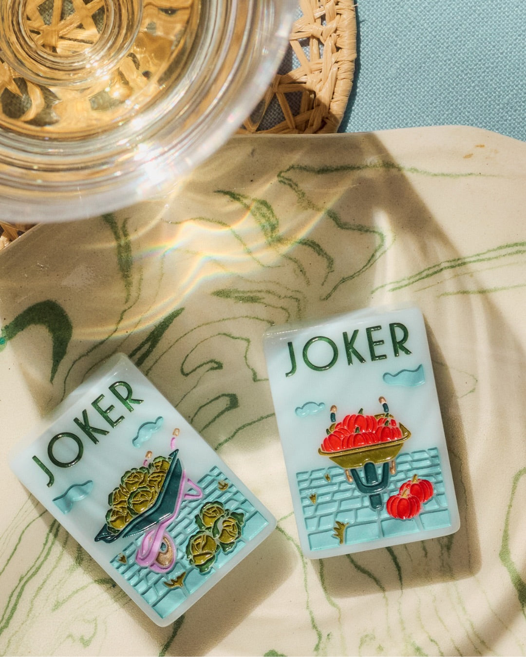 Two Joker Mahjong rectangular tiles with illustrated designs, placed on a light marbled surface with a glass and woven mat partially visible in a styled setting at The Queen's Mahjery in Charlotte NC.