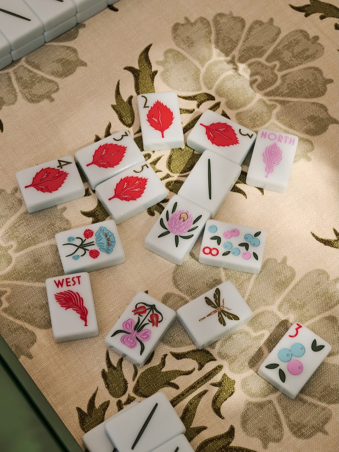 A set of Mahjong tiles with floral and feather designs scattered on a beige floral-patterned fabric surface at The Queen's Mahjery Charlotte NC's Mahjong Studio.