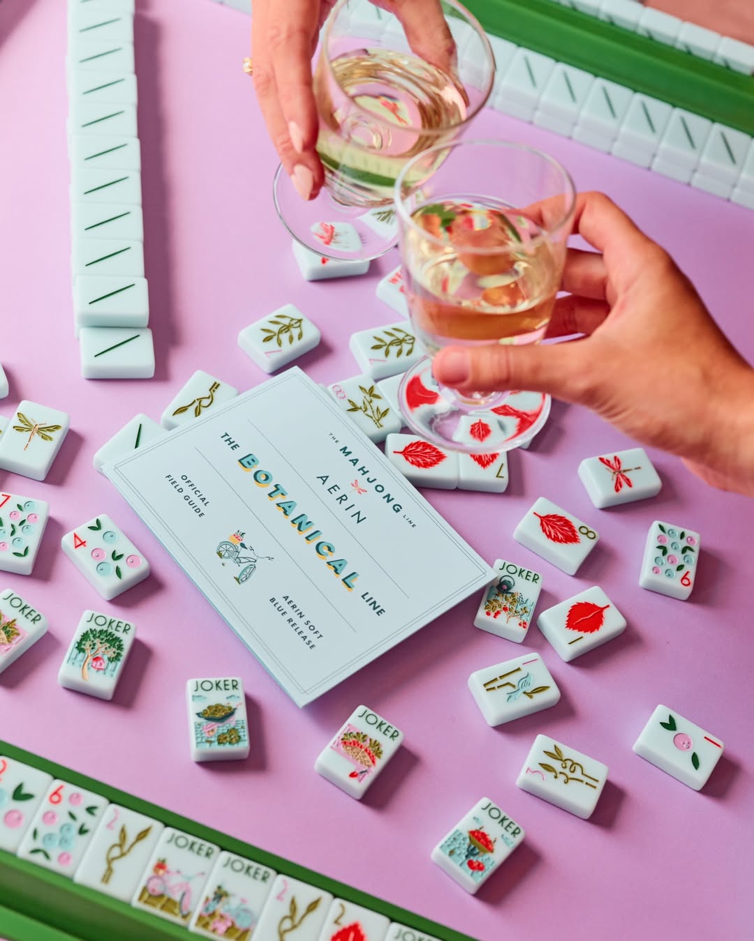 A styled flat-lay of a mahjong game set on a pink surface, with hands holding drinking glasses over the tiles. The set includes white tiles with colorful illustrations and a light blue instruction card reading 'THE BOTANICAL LINE' and 'The Mahjong Line' at The Queen's Mahjery in Charlotte NC.